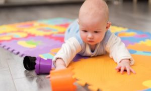  A baby is laying on their tummy on a colorful play mat stretching to reach a small cup.