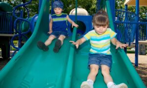 Two preschool age children ride down the double slide on a small playground.