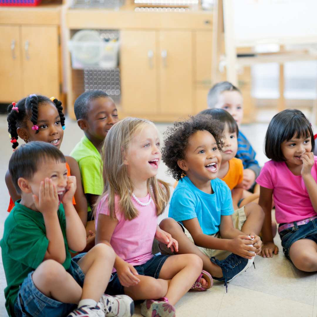 Group of preschool children in a brightly lit classroom sit on the floor for circle time.