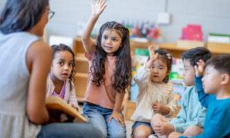 A female Montessori teacher sits in front of a small group of children as she reads them a story.