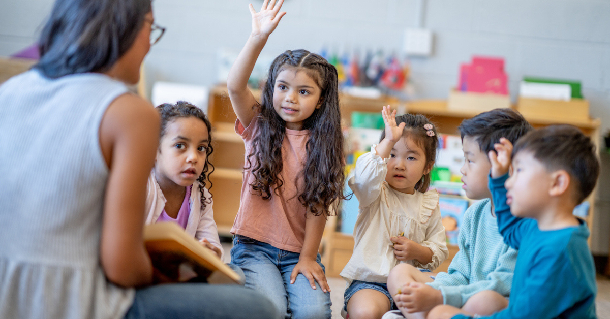 A female Montessori teacher sits in front of a small group of children as she reads them a story.