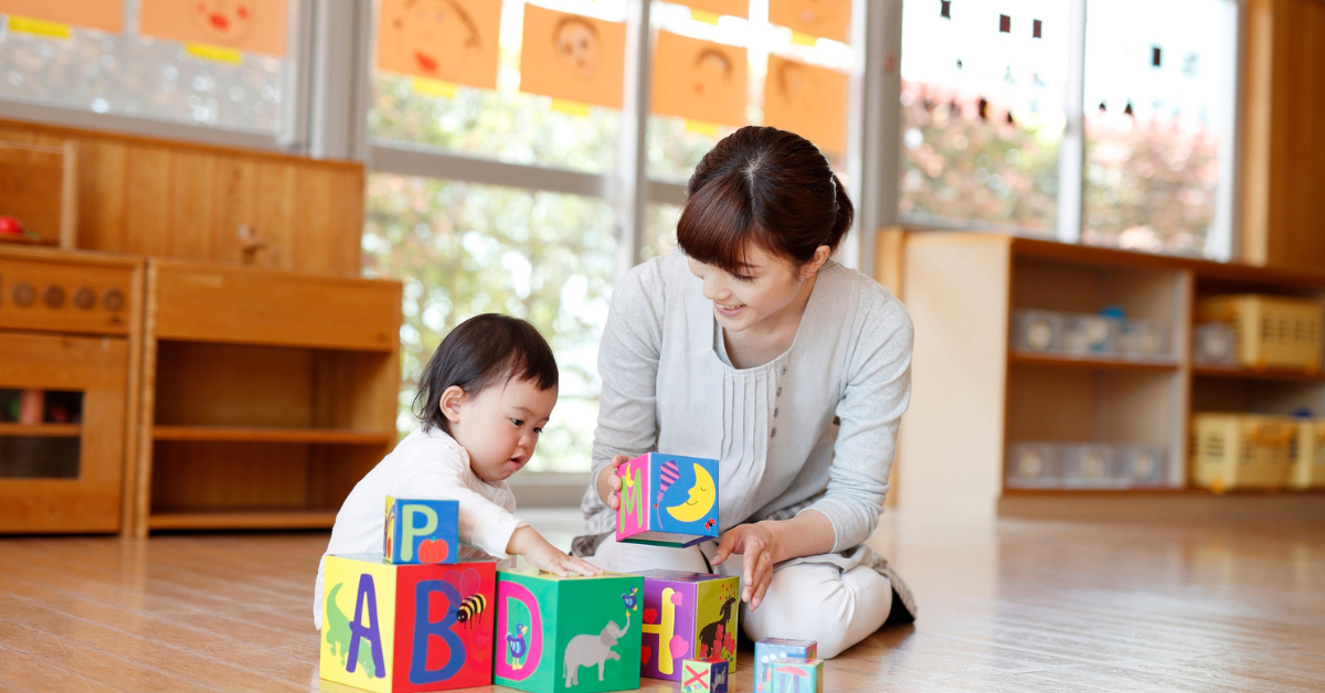 a child care provider and child sit on the wooden floor in a home day care.