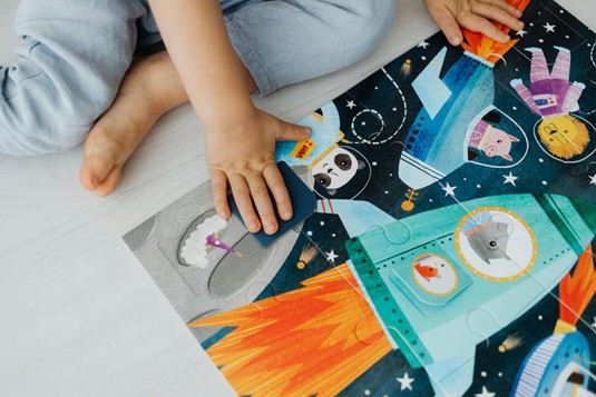 Child completing a colorful space-themed puzzle on the floor, featuring cartoon animals in a rocket ship.